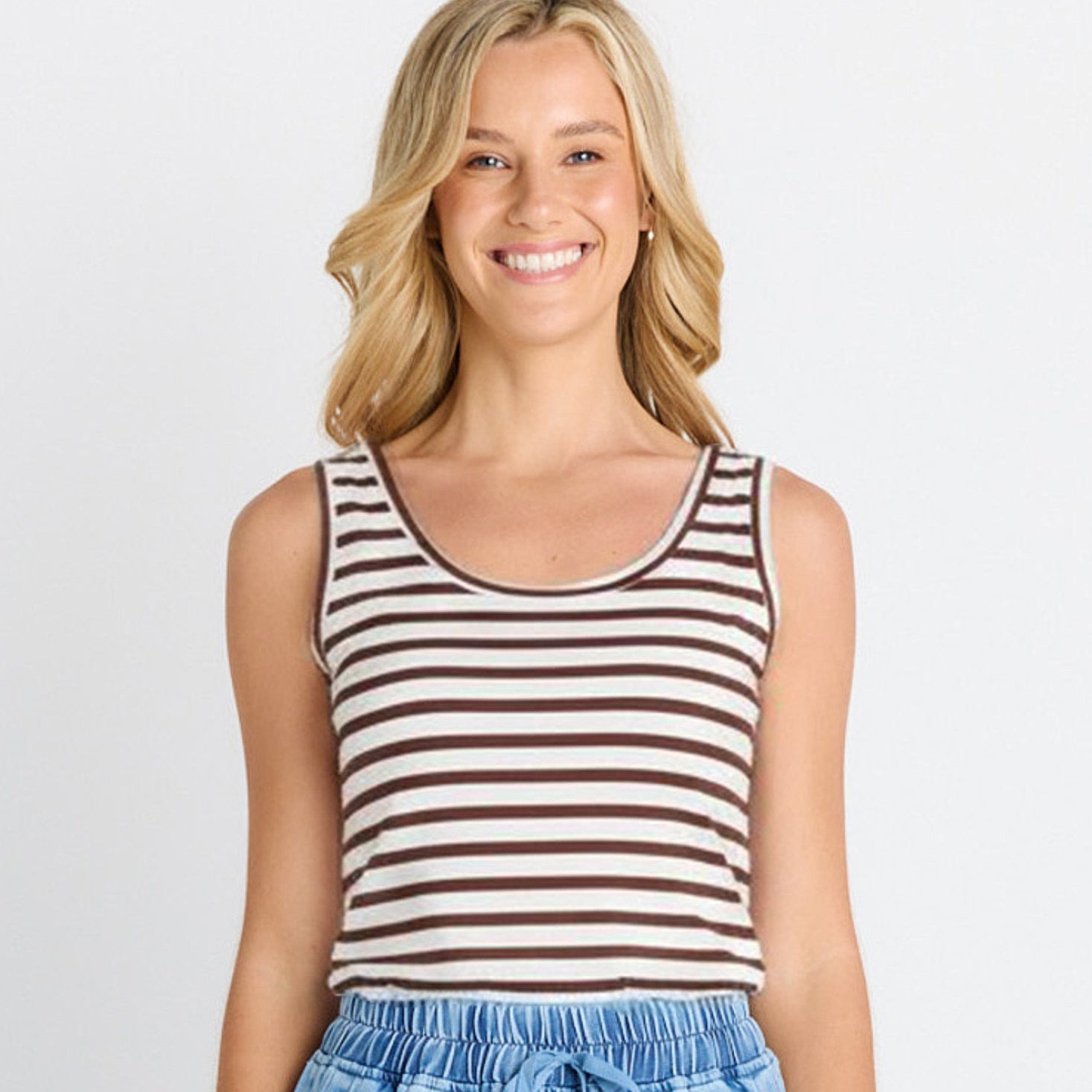 A smiling blonde woman wearing a brown and white horizontally striped tank top with light blue denim shorts, photographed against a light gray studio background.