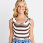 A smiling blonde woman wearing a brown and white horizontally striped tank top with light blue denim shorts, photographed against a light gray studio background.