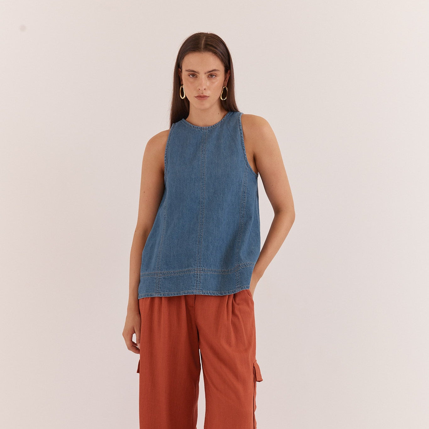 A woman stands against a plain off-white studio background wearing a sleeveless blue denim top and rust-orange wide-leg pants with side cargo pockets.