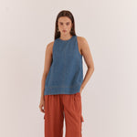 A woman stands against a plain off-white studio background wearing a sleeveless blue denim top and rust-orange wide-leg pants with side cargo pockets.